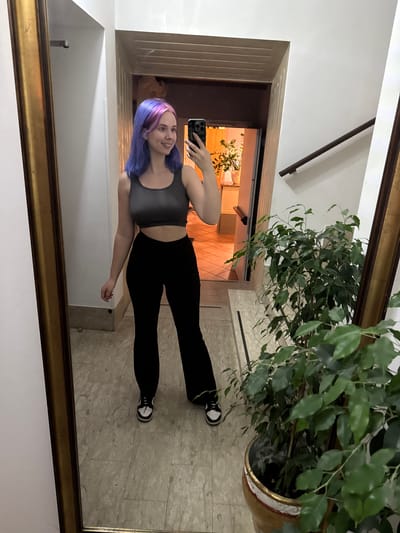 A young woman with purple hair is reflected in a large, ornate mirror. She is holding a smartphone up to take a selfie. She is wearing a gray top, black leggings, and black and white sneakers. Behind her in the reflection is a hallway with a tile floor. There is a large potted plant in the upper right corner of the mirror's reflection. Based on the interior architecture and the presence of a large ficus plant, the location is likely an apartment or home interior in Villafranca di Verona, Italy.