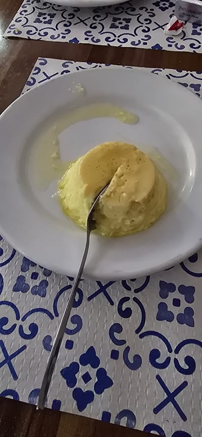 A close-up, overhead shot shows a single serving of creme caramel, also known as flan, on a white plate. The dessert is a light yellow, dome-shaped custard with a liquid caramel sauce pooled around its base. A silver spoon is partially submerged in the dessert, with a small portion already removed. The plate rests on a blue and white patterned placemat, which is on a dark wooden table. The placemat has a repeating pattern of stylized floral elements and geometric shapes. The background is slightly blurred, but the hint of a red and white object, possibly a cigarette pack, can be seen at the top right corner. This image does not contain enough information to determine its location in Venta de la Romera, Spain.