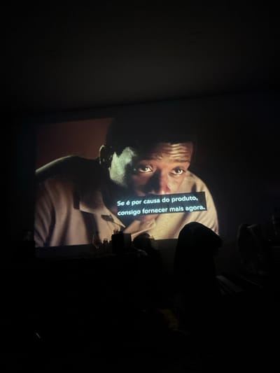 This image depicts a dark room in Pedrouços, Portugal, illuminated by a projected film or television scene. The projection, occupying the upper two-thirds of the frame, displays a close-up of a dark-skinned male actor with a serious expression, looking upwards and to the right. He is dressed in a light-colored collared shirt. Portuguese subtitles are superimposed on the screen, reading: "Se é por causa do produto, consigo fornecer mais agora." Below the projected image, a dark, flat surface is visible, bearing indistinct objects including what appears to be a small bottle and a glass. In the lower right, dark silhouettes of at least two individuals, presumably seated viewers, are discernible, indicating an audience engaged with the projected content. The environment suggests an indoor, domestic setting with minimal ambient light.