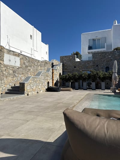 The image displays an outdoor leisure area, characteristic of Mykonos, Greece, under a clear, cloudless blue sky. The foreground on the right shows the upper portion of a brown, textured outdoor lounge chair. The ground consists of large, light grey rectangular paving tiles, extending to a turquoise swimming pool on the right midground.
Architecturally, the scene features traditional Cycladic elements: whitewashed buildings atop natural stone walls. On the left, a large whitewashed structure surmounts a substantial dry-stacked stone wall. Integrated into this wall are grey steps with a distinct wavy grey handrail, illuminated by small embedded lights. A window with closed white louvered shutters is visible within the stone wall. In the midground, another stone wall supports a separate whitewashed building on the right, which features a balcony with light blue-grey shutters and a single dark chair. Below this building, a row of five small, manicured trees in tall grey planters stands along the paved area. To the far right, a folded light-colored patio umbrella is situated by the pool's edge. Two dark, beanbag-style loungers are positioned on the paved area at the base of the left stone wall. No individuals are present in the scene.
