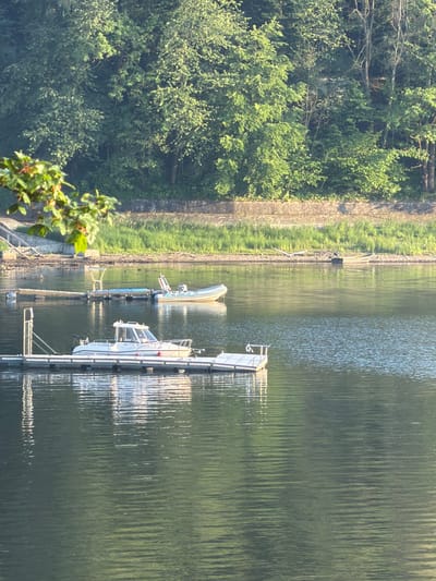 The image shows a body of water with several boats docked. In the foreground, a white motorboat is tied to a floating dock. Further out on the water, there is another smaller boat, possibly a dinghy, and a larger inflatable boat with a motor. The water is calm with ripples reflecting the sunlight and the surrounding trees. In the background, a lush green forest borders the water, with a stone embankment visible along the shoreline. There are no people visible in the image. The scene is set in Lipie, Poland.