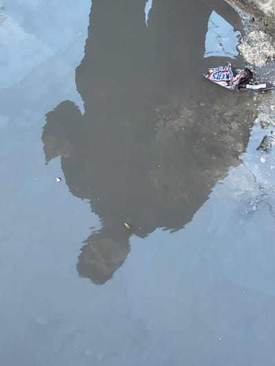 The scene depicts a close-up view of a shallow, still body of turbid greyish-brown water located in the city of Qudsaiya, Syria. A dark, inverted reflection of a human figure is prominently visible, showing the upper torso and head in the central and left portions of the water's surface. The reflection indicates an individual standing above and observing the water; specific facial features are not discernible. Along the upper right edge of the water, a crumpled "SNICKERS" candy wrapper is partially submerged, resting against an irregular, dark bank composed of earth and scattered debris. Minor fragments of debris are visible within the water. A small, light-colored, elongated object floats near the reflected head. The water's surface exhibits subtle reflections of an overhead light source.