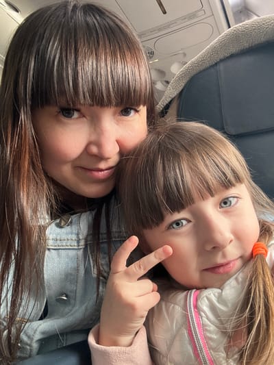 A close-up, tilted portrait features two females, an adult and a child, sharing a tender moment, likely within an airplane cabin. Both subjects are looking towards the viewer, suggesting a selfie.

The adult, positioned in the lower half of the frame, has straight, dark brown hair with bangs that cover her forehead. Her eyes are a striking blue-gray, and she wears a subtle, soft expression with a slight smile. A light blue denim jacket is visible on her shoulder and chest.

The child, positioned slightly above and to the right of the adult, has lighter brown hair with bangs, and bright blue eyes that are wide open. Her hair is partially gathered into a ponytail secured with a bright orange elastic tie. She is wearing a shiny, light-colored puffer jacket with a distinct pink and white striped trim along what appears to be a zipper line. The child is playfully making a "peace sign" or "V" gesture with her right hand.

The background clearly indicates an airplane interior. A grey, textured seat headrest is visible in the upper left, while the overhead panel with air vents and lights can be seen in the upper left and center background. The subjects appear relaxed and engaged, with the child showing a playful demeanor. There is no visible text in the image. The provided location context is Kitsjanovo, Russia.