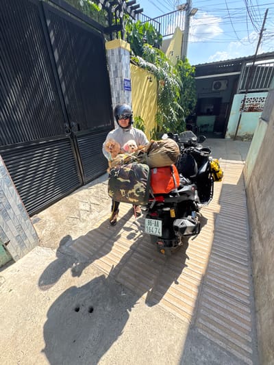 A person wearing a light-colored jacket and helmet stands by a black metal gate, holding two small dogs, one tan and one white, in front of a motorcycle loaded with luggage. The motorcycle is parked on a sidewalk with textured concrete. The background shows a yellow wall with green foliage, a building with an air conditioning unit, and a blue sky with clouds. A license plate on the motorcycle reads "86-AA 164.74". The scene appears to be in Binh Thuan Ward, Vietnam, suggested by the license plate format.