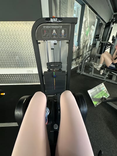The image shows a first-person perspective of someone using a hip abductor/adductor machine in a gym. The machine is black and has a weight stack. The user's legs, wearing light-colored leggings and athletic shoes, are visible in the foreground. In the background, another person is visible using a leg press machine. The gym environment features gym equipment, a textured wall, and large windows. The gym is identified as being located in Carlow, Ireland.