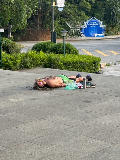 In the city of Hy Cương, Vietnam, a person is lying on their stomach on a paved walkway. The individual is wearing a green shirt and black shorts, with blue socks and black shoes. A colorful headscarf is on their head, and dirt and debris are scattered around them. Next to the person are a green plastic bag, some clothing items, a water bottle, and a shoe. In the background, there is lush green vegetation, a street with yellow markings, a lamppost, and a large blue sign with Vietnamese text and the VNPT logo. The sign appears to be promoting an event. The scene is illuminated by daylight, casting shadows on the ground.