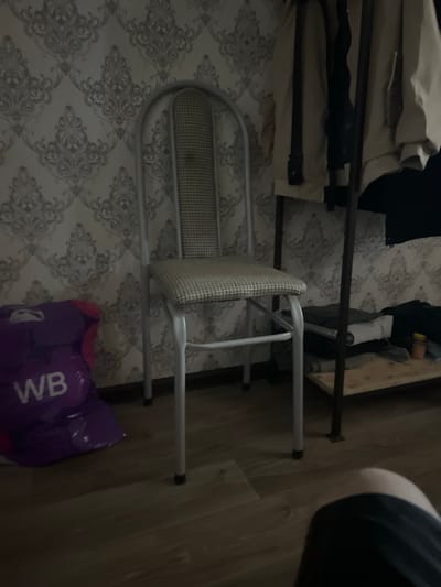 The image shows an indoor scene with a metal folding chair in the foreground. The chair has a woven seat and backrest and is positioned on a patterned floor. Behind the chair, there is a dark-colored metal frame, possibly part of a bed or shelving unit, with some fabric draped over it. Further back, a portion of a wooden surface is visible, which could be furniture or part of a room's structure. On the floor to the right of the chair, a purple plastic bag with white lettering is visible. The lighting in the image is dim, suggesting it might be evening or the room is not well-lit. The overall impression is of a simple, perhaps utilitarian, living space. There is no indication of any people or specific activities taking place. The visible text on the bag appears to be "WB".