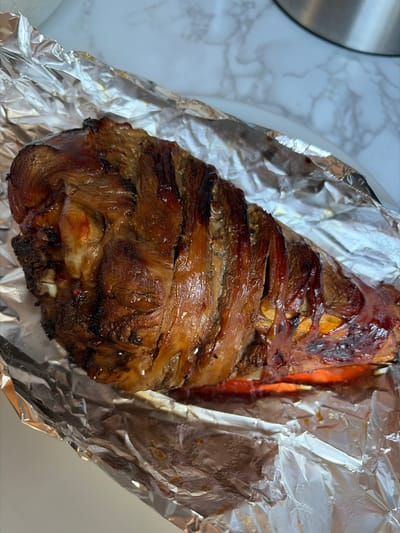 A close-up, overhead shot captures a succulent, roasted turkey leg wrapped in crinkled aluminum foil. The turkey skin is a deep, glistening brown, with hints of charring in places, suggesting a rich, savory flavor. The foil, also shiny, partially obscures the turkey, adding a rustic presentation. The background is a soft, out-of-focus marble countertop, providing a clean and simple setting. No people or discernible location in Göttingen, Germany are visible.