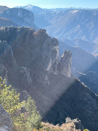 The image presents a rugged mountainous landscape in the vicinity of Klobuk, Bosnia and Herzegovina. The foreground consists of a rocky terrain partially covered by illuminated green pine trees and areas of dry, brownish brush. A distinct, sharp diagonal shadow traverses the lower right quadrant of the image, obscuring the terrain within its bounds. The midground features a prominent formation of jagged, grey rock cliffs and spires, characterized by steep faces and varying light exposure, creating areas of highlight and deep shadow. These rock formations descend into a vast valley or canyon. The background reveals multiple layers of progressively lighter blue-hued mountain ranges, indicative of atmospheric perspective and distance. The most distant peaks are capped with visible patches of snow under a clear, light blue sky. No people, animals, man-made objects, or specific actions/interactions are present within the frame.