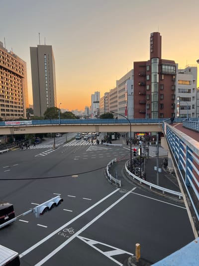 An elevated view captures a Japanese city street at dusk. Buildings line the left side, silhouetted against an orange sky. A roadway below features multiple lanes, white markings, and a designated bicycle lane. Traffic signals are visible. A bridge or elevated structure forms the top of the frame, with a blue and white barrier. Bicycles are parked on the edge of the sidewalk.