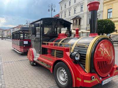 This is a medium shot, eye-level photograph of a red and black miniature train parked on a cobblestone plaza. The train, labeled "NEUSOHL EXPRESS," appears to be a tourist attraction, with multiple carriages behind the engine. The engine is brightly decorated with gold accents and features a large, round grill at the front, reminiscent of a steam engine's boiler. A headlight is visible on the side of the engine. The carriages are mostly black with red trim and have a series of open windows.
In the background, there are buildings with classical architecture, suggesting an urban setting. The sky is overcast, indicating a cloudy day. Several people are visible in the distance, walking on the plaza and sitting at outdoor tables, some under umbrellas. One of the building facades has signage that includes "Coca-Cola." The license plate on the train reads "AA 481GX." The overall impression is a bright, sunny day in a historic European city, with a funicular train ready to transport visitors.