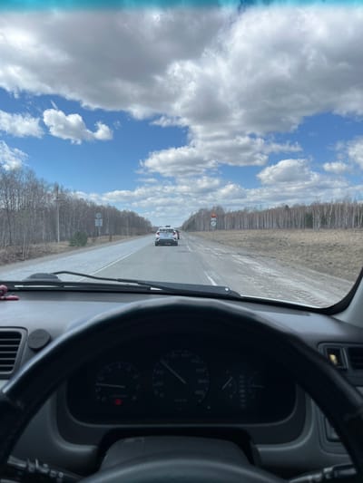 The image shows a view from inside a vehicle, likely a car, traveling on a straight road in Mar'evka, Russia. A white vehicle is traveling ahead. On the left and right sides of the road are sparsely wooded areas. The sky above is blue with scattered white clouds. Part of the steering wheel and dashboard is visible in the foreground.