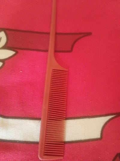 The image displays a close-up, top-down view of a red comb resting on a red patterned fabric surface. The comb is positioned vertically, with its teeth facing downwards. The fabric has a textured appearance and features a repeating pattern of dark red outlines and white shapes. The lighting appears to be artificial, casting subtle shadows. There is no indication of the location being Majemu, Nigeria, or any specific urban environment. The scene solely consists of the comb and the fabric.