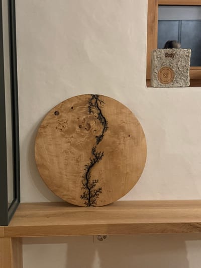 A round wooden cutting board with a black lightning bolt design is placed on a wooden shelf in a home in Celerina/Schlarigna, Switzerland (latitude: 46.514, longitude: 9.859). The board is resting on a light-colored wooden shelf that is part of a shelving unit against a white wall. A small white rectangular object with a black circle and the word "Body" inscribed on it is sitting on a shelf above the wooden board, partially obscured by the corner of a black-framed window.  The cutting board is positioned in the middle of the shelf, with the bottom edge of the board even with the front edge of the shelf.  The shelf is partially obscured by the shadow of a window. The object on the upper shelf is partially obscured by the window frame.  The wall and shelf appear to be constructed of a light-colored wood, and the cutting board is made of a darker wood.  The overall scene is characterized by minimalist design elements and muted tones.
