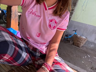 The image presents a close-up, eye-level perspective of an individual seated outdoors in Desa Batu Layar, Indonesia. The subject, largely cropped at the shoulders and upper thighs, wears a light pink short-sleeved shirt with "Vérco" in script on the chest, a pink and white shield-shaped emblem on the upper right, and a subtle graphic below the text. Paired with this is a pair of long pants featuring a red, dark blue, and white plaid pattern. The individual's right arm is visible, adorned with multiple colorful string and bead bracelets on the wrist. They are seated on a slatted, light-colored surface, likely bamboo. In the background, to the left of the subject, a dark, weathered wooden post is visible next to a partially obscured blue scooter or motorcycle and some colored bags (blue and purple). To the right, a light green wall, with a grey concrete base, displays faint white markings or drawings. Leaning against this wall is a small blue balance bike, bearing the text "IÕIO" on its frame. Adjacent to the bike, a large, brown woven basket, appearing to hold laundry or textiles, rests on its side on the uneven, light-brown dirt ground. Strong sunlight illuminates the scene, casting shadows and indicating an outdoor environment.