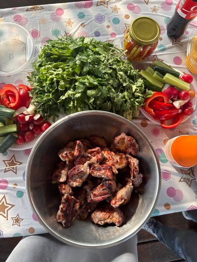 The image shows a spread of food on a table with a patterned tablecloth. A large metal bowl is filled with grilled meat, possibly shashlik. There is a large bunch of cilantro next to the bowl. Plates of sliced vegetables, including cucumbers, tomatoes, radishes, and red bell peppers, are also on the table. A jar of pickled vegetables, an orange cup, and a bottle of Coca-Cola are visible. The scene appears to be a gathering or picnic of some sort. The location information indicates that this image was likely taken in or near the city of Кузнецово, Russia. A person is partially visible in the upper right of the image. The setting is outdoors, as indicated by the lighting.