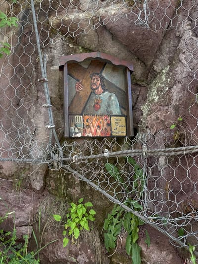 This is a photograph of a painting depicting Jesus Christ carrying the cross. The painting is mounted in a weathered, dark-colored frame and is affixed to a rocky hillside, partially obscured by a wire mesh fence and metal cables. The painting shows Jesus from the chest up, with a crown of thorns on his head and his gaze directed downwards. He is carrying a large, brown cross. To the right of Jesus's head, there are smaller figures painted in an abstract, colorful style, possibly representing souls or a depiction of suffering. Above these figures, in a yellow box, is text in a foreign language. The text appears to read "Prega per l'anes de Purgatuere". The background of the painting is a mix of blues and browns. The surrounding setting is natural, with reddish-brown rocks and patches of green vegetation growing around the painting and the wire mesh. Sunlight illuminates the scene, suggesting it is daytime. The overall impression is one of religious devotion and a sense of place on a mountainside in Urtijëi - St. Ulrich in Gröden - Ortisei, Italy.