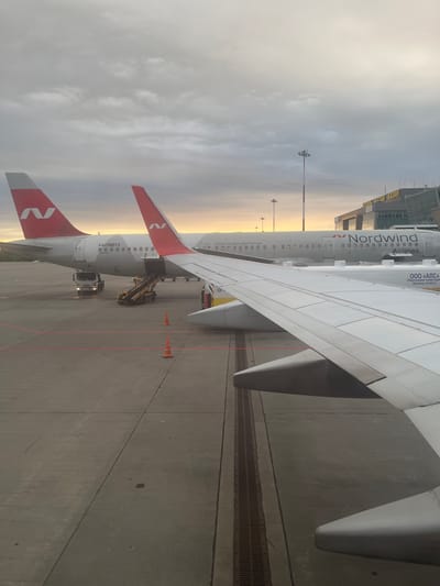 The image captures a scene at an airport with a Nordwind Airlines airplane parked on the tarmac. The airplane's wing extends prominently into the foreground, with its tail fins, featuring the airline's red and white logo, visible on the left side. The aircraft's fuselage bears the registration "RA-73273" and the airline's name, "Nordwind Airlines," along with its website address.

On the ground, a baggage loader is positioned next to an open cargo door of the aircraft, indicating that baggage handling or loading is in progress. A white truck is also parked nearby. Orange traffic cones are placed on the tarmac, likely to demarcate areas or guide vehicles. In the background, an airport terminal building is visible, with a glass facade and what appears to be a skybridge. Lampposts stand tall against the sky.

The sky is overcast with hints of soft sunlight breaking through the clouds, suggesting either early morning or late afternoon. The overall lighting and atmosphere evoke a sense of an ordinary airport operation. The location is identified as Stolbishche, Russia. The visible text on the airplane includes company names and phrases related to service.