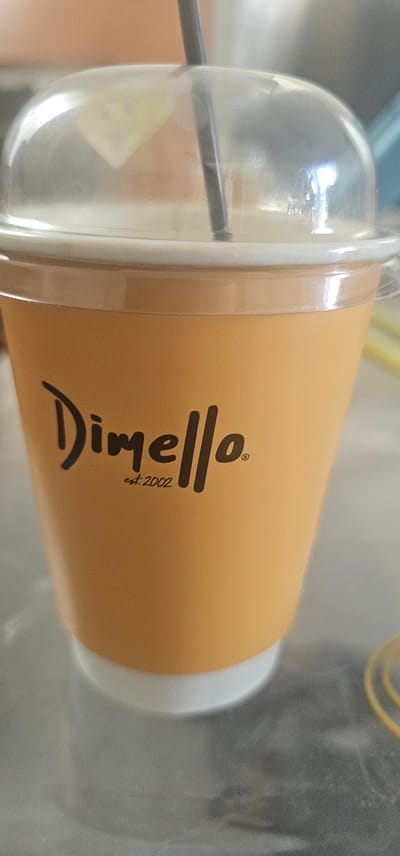 The image shows a close-up view of a takeaway coffee cup with a domed lid and a black straw. The cup is orange and has the logo "Dimello" printed in dark brown, with "est. 2002" below it. The lid is clear plastic, and the drink inside appears light brown. The cup is placed on a grey surface, possibly a table. In the background, there is a blurred out pinkish-orange wall and a sliver of a blue object. The overall lighting suggests an indoor setting. There is no indication of the location in Paralimni, Cyprus, as the image does not contain any identifiable landmarks or textual clues to that effect.
