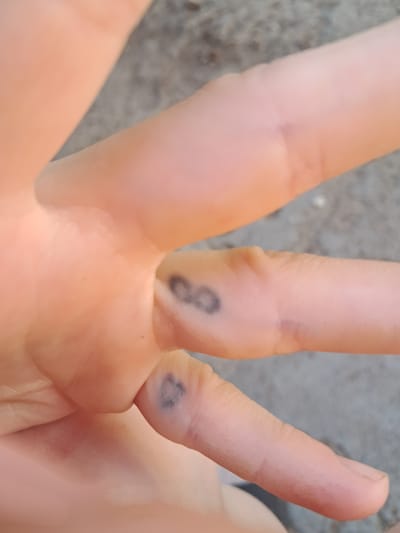 The image shows a close-up view of a person's hand. There are two small, dark blue tattoos on the fingers. The tattoo on the middle finger appears to be an infinity symbol. The tattoo on the ring finger is partially obscured but seems to be a similar shape. The skin is light-toned. The background is out of focus and appears to be a rough, grey surface, possibly concrete or asphalt, suggesting an outdoor urban or rural setting. There is no indication of the location being Chobruchi, Moldova.