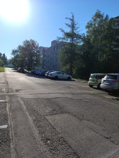 A low-angle, sideways view captures a row of parked cars on an asphalt surface under a clear blue sky. The sun is shining brightly, casting long shadows and creating a lens flare on the left side of the image. To the left, a multi-story building with many windows stands behind a dense canopy of green trees. Another building, also with many windows, is visible further in the distance. The asphalt parking lot has painted white lines marking parking spaces, and there are visible cracks and patches on the surface. Among the parked vehicles, a light green car is prominent in the foreground, followed by a silver car and a dark-colored SUV. Further down the line, a white car and a dark red car are also visible. The scene appears to be a typical residential or public parking area on a sunny day. The environment suggests an urban or suburban setting.