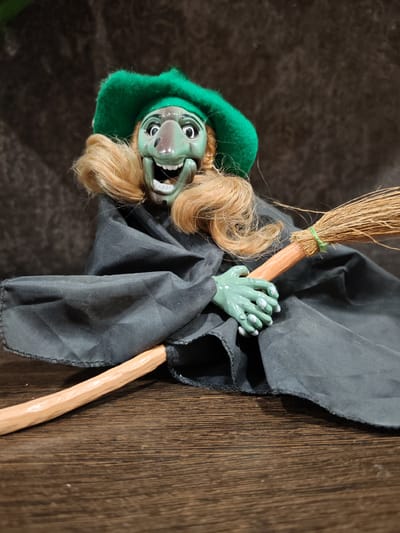 The image shows a close-up, eye-level, indoor shot of a doll dressed as a witch. The doll has a green face with wide, black-rimmed eyes and an open mouth showing white teeth. It is wearing a green felt hat and a black cloak. The doll is holding a wooden broomstick with straw bristles. The broomstick is angled across the doll's lap. The doll is positioned on a dark wooden surface. The background is a dark, textured fabric. The location in the city of Buy, Russia, is not discernible from the image.