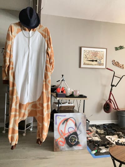 The image depicts an indoor scene, possibly a residential room or a studio space. A giraffe-patterned onesie with a white chest panel is hanging from an unseen support, with a dark fedora hat placed on top of the onesie's hood. A silver chain necklace is draped around the neck area of the onesie.
To the right, a small red scooter with a black handlebar and red grips is leaning against the wall. A framed picture hangs on the wall above the scooter, depicting a colorful, abstract design with geometric shapes and figures. Two decorative wooden pieces with letters are also mounted on the wall, one reading "SEKA".
In the mid-ground, a black metal table with a wooden top holds a black tripod, a white cup, and a small glass with a pink pattern. To the right of the table, a gray bucket is visible.
In the foreground, a square canvas with spray-painted abstract art is placed on the floor. The art features orange, purple, and black spray paint. The floor is cluttered with various items, including what appear to be electronic components, wires, and other debris, some of which are arranged on a black mat outlined in blue tape. A round wooden table is partially visible in the lower right corner. The room has light-colored walls and a concrete or linoleum floor. The overall impression is one of an artistic or somewhat disorganized personal space.
This image was captured in Round Rock, United States of America.