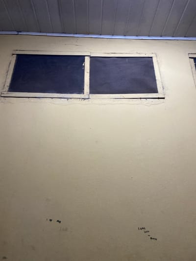 The image shows a portion of an exterior wall, likely a building or house in Sabo, Nigeria. The wall is light beige with a rough, textured surface. A gray siding is visible in the left of the frame. There is an inset window. The window frame is off-white. The window is divided into two separate dark planes. There are faint blue markings are present on the wall to the upper right. There are also faint blue markings on the lower right of the frame.