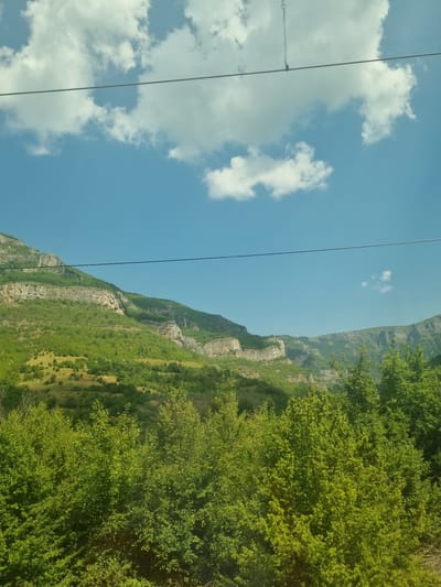 This is a landscape photograph taken from a slightly elevated vantage point, looking towards a mountain range under a partly cloudy sky. In the foreground, there is a dense growth of lush green trees and foliage. The middle ground features a sloping hillside covered in trees, transitioning into a rocky, layered mountain face. The sky is blue with scattered white cumulus clouds. A thin, dark horizontal line, possibly a power line or cable, stretches across the upper portion of the image. The image appears to be taken in a natural, mountainous region, likely Ochindol, Bulgaria, given the context. There are no people or discernable actions taking place.