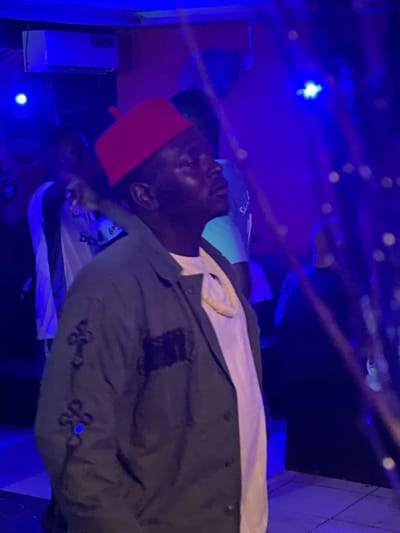 In a dimly lit interior, likely a club or event space in Durumi, Nigeria, bathed in vibrant blue and purple lighting, a person wearing a red cap and a dark jacket is the central subject. Their head is tilted upwards, eyes closed, and their expression suggests a moment of reflection or enjoyment. The jacket has intricate, dark embellishments on the sleeves and a patch on the chest. A light-colored necklace or scarf is visible around their neck, peeking out from a white shirt. In the background, other individuals are present, partially obscured by the low lighting and the subject's presence. An air conditioning unit is mounted on the wall above and to the left. The environment appears to be a public gathering space, possibly a night club, with the characteristic mood lighting and a sense of activity.