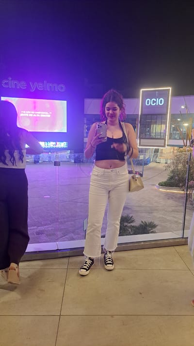 Two women are standing in front of a glass barrier in Alcorcón, Spain. The woman on the right is facing the camera and holding a smartphone. She is wearing a black crop top, white jeans, and black Converse sneakers. She has curly, dark hair with purple highlights and a small purse hanging from her shoulder. The woman on the left is partially visible and is facing away from the camera. She is wearing a white off-the-shoulder top and dark pants. Behind the women, a large screen displays "cine yelmo" and text in Spanish. Another sign in the background reads "OCIO." The environment appears to be an outdoor entertainment or shopping complex with modern architecture, bathed in purple and white lighting.