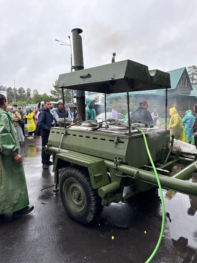 In Местечко Раифа, Russia, on a cloudy day, a group of people are gathered around a large, olive-drab, military-style field kitchen mounted on a trailer. The field kitchen has multiple large pots on top, with steam rising from them. A green hose is connected to the side of the kitchen. Several individuals are wearing raincoats, suggesting inclement weather. The people are standing on a wet, asphalt surface, with reflections visible. In the background, there is a wooden building with a green roof, trees, and some vehicles. The overall scene appears to be an outdoor event or gathering where food is being prepared and served from the mobile kitchen.