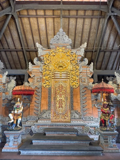 This image displays the elaborately decorated entrance of a traditional temple structure in Batuan, Indonesia. A central, double-paneled wooden door, intricately patterned in red and gold with floral and geometric designs, is framed by ornate golden carvings. Above the door, a prominent golden carving of a fierce-faced mythological figure, likely a Boma or Kala head, is positioned. This central feature is integrated into a reddish-brown stone wall, extensively embellished with grey volcanic stone carvings depicting mythological figures, foliage, and architectural motifs.

Flanking the entrance are two large, polychrome Dvarapala guardian statues, each standing on a multi-tiered stone pedestal. The left guardian statue is light-skinned with a green face, holding a dark club in one hand and a red ceremonial umbrella (payung) in the other. The right guardian statue is red-skinned with a red face, holding a dark club and a red ceremonial umbrella. Both figures feature prominent fangs and wide, painted eyes.

In front of the door, a set of three dark grey stone steps ascends to a platform. Overhead, a high-pitched roof structure is visible, with its underside composed of light-brown woven material, likely palm or bamboo leaves, supported by exposed dark wooden beams and a truss system. No human subjects are present in the scene.