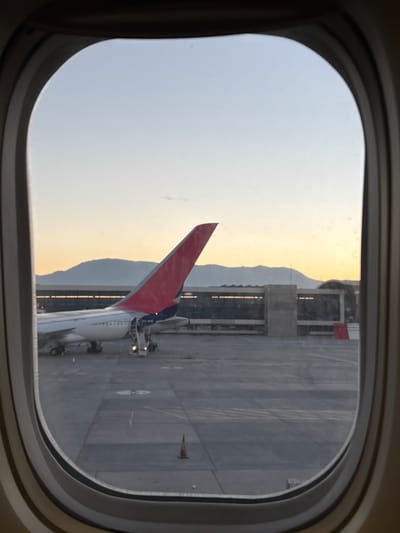 The image is taken from inside an airplane, looking out of a window. The airplane is on the ground, as evidenced by the view of the airport apron. A building is next to the plane. The vertical stabilizer of the airplane is visible and has a red upper portion. The airport apron is a gray concrete surface with various markings and equipment. In the distance, a mountain range is present, with a faint golden hue likely indicating either sunrise or sunset. The sky is pale. The location is likely in Pudahuel, Chile.