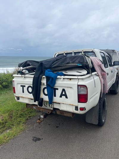 A white, dual-cab Toyota utility truck is parked on an asphalt surface in Wombarra, Australia, positioned next to a grassy embankment overlooking the ocean. The sky above is overcast with grey clouds. The vehicle's rear tailgate displays "TOYOTA" in large black lettering and exhibits rust and wear. A New South Wales license plate, "92-VZ", is affixed below the tailgate, above a visible tow hitch. The truck bed contains multiple surfboards, some covered in black board bags, along with at least two uncovered white surfboards. A black wetsuit and a blue garment, appearing to be another wetsuit or rash guard, hang draped over the tailgate. A pink garment, possibly a towel or poncho, hangs from the passenger side of the truck bed. A wooden tool with a black attachment, consistent with a surfboard wax remover, hangs near the black wetsuit. The truck is equipped with large, off-road tires, with "BFGoodrich" visible on the sidewalls. To the left of the vehicle, dense green coastal vegetation lines the edge of the road, beyond which the ocean is visible with breaking waves. No individuals are present in the scene.
