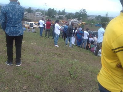 A group of people are gathered on a grassy hill. Several yellow and white vans are parked in the background. The people are dressed in a variety of casual clothing, including blue jeans, white shirts, and patterned dresses. Some of the people are standing, while others are sitting on the grass. There are buildings and trees visible in the distance. The scene appears to be an outdoor gathering or event.