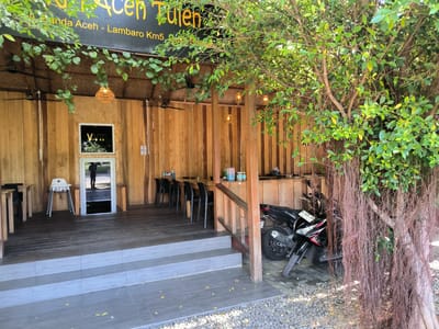 The image shows the exterior of a building in Santan, Indonesia. The structure is primarily constructed with wooden planks. A sign above the entrance reads "Bunda Acen Tulen" and "Inda Aceh - Lambaro Km5". The entrance features three steps leading to a wooden deck. Inside, there are tables and chairs arranged. A high chair is visible near the entrance. A motorcycle is parked to the right of the building, partially obscured by a tree with hanging roots. The area around the base of the steps is covered in small pebbles.