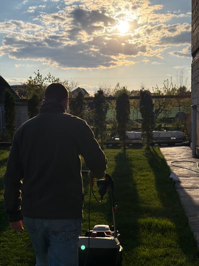 A man is mowing a lawn in a backyard setting. The sun is setting or rising, visible through the clouds in the sky. The man is operating a lawnmower. The location is likely Kurylivka, Ukraine.