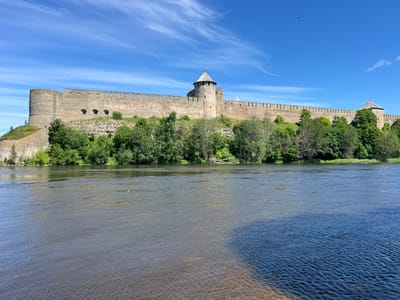This media file captures a scenic view of the Narva Castle, a medieval fortress located in Narva, Estonia. The castle's imposing stone walls and towers stand prominently atop a rocky hill, bathed in the bright light of a clear, sunny day. Lush green trees and vegetation surround the base of the hill, creating a stark contrast with the ancient stone architecture.
The foreground is dominated by a wide river, its surface shimmering with ripples. The water appears to be a mix of lighter, perhaps shallower, sandy areas and deeper, darker blue sections. The distinct color change in the river might indicate a confluence of waterways or variations in the riverbed.
The sky above is a vibrant blue, dotted with wispy white clouds. Numerous small, dark specks are visible in the sky, likely birds in flight, adding a sense of life and movement to the otherwise static landscape. The overall atmosphere is serene and majestic, highlighting the historical significance and natural beauty of the location. The time of day appears to be midday, given the strong sunlight and shadows. There is no visible text in the image.