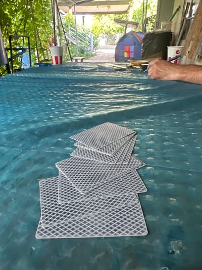 A person's arm and hand are visible on the left side of the frame, reaching towards a stack of playing cards fanned out on a dark teal, textured surface. The cards have a checkered back pattern. The background shows a blurred scene of greenery and some structures, suggesting an outdoor or semi-outdoor setting. A red and white object, possibly a sign or container, is visible in the mid-ground on the left. The lighting suggests it is daytime.