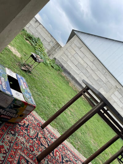 In Nartan, Russia, an outdoor scene unfolds with a focus on a corner of what appears to be a yard or an enclosed outdoor space. A large, partially opened cardboard box, bearing markings that suggest it contains bananas and the brand "MATHIAS", rests on a patterned rug. Nearby, a metal barbecue grill stands on the grass. In the background, a tall wall constructed of concrete blocks forms a boundary, with green vegetation growing along its base. Beyond this wall, a building with a corrugated metal roof is visible, angled against a cloudy sky. The overall impression is one of an unfinished or utilitarian outdoor area.