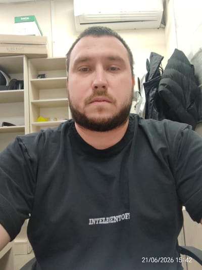 The image is a selfie of a man with a beard, wearing a black t-shirt with white lettering. He is in an office or storage area, with shelves filled with boxes and other items behind him. On the right side of the image, there are several dark-colored jackets hanging on a rack. The lighting appears to be artificial. A date and time stamp in the bottom right corner reads "21/06/2026 15:42". The scene appears to be indoors, and the overall environment suggests a functional workspace.