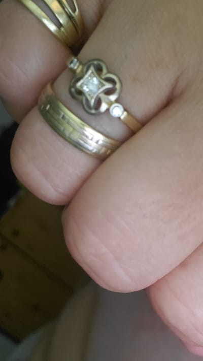 The image shows a close-up of a person's hand wearing three gold rings. The ring closest to the viewer has a band with horizontal lines and two small circular stones on either side. Above this ring is another ring with a clover-like design in the center, featuring a square-shaped diamond, and two smaller circular stones on the band. The third ring, partially visible at the top of the image, is a wider gold band with a split design. The background is out of focus, showing a dark, possibly wooden surface and some indistinct shapes. There are no discernible landmarks or indications of a specific location within the city of Sasbach, Germany, in this image.