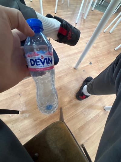 A first-person perspective shot shows a hand holding a bottle of DEVIN mineral water. The bottle has a blue cap and a label with pink mountains and the DEVIN logo. The background shows a wooden floor with several white chairs and a dark fabric object, possibly a bag or clothing. The scene appears to be indoors, possibly in a classroom or common area. There are no visible indications of the city of Satovcha, Bulgaria.