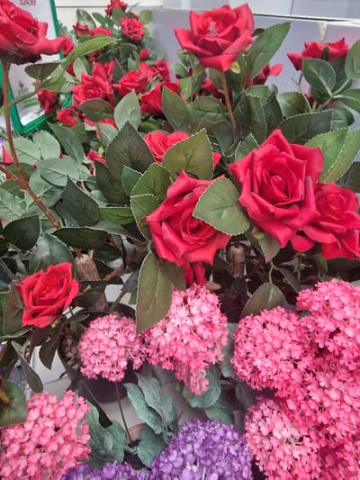 The image shows a close-up of an artificial floral arrangement. Dominating the foreground and midground are several large, vibrant red roses with green leaves. The roses appear to be made of fabric or plastic, with defined petals and realistic-looking veins on the leaves. Interspersed with the roses are clusters of smaller pink flowers, resembling hydrangeas, and some purple flowers. The arrangement is in a dark pot, which is partially visible. In the upper left corner, a green sign with white text is visible, partially obscuring some of the flowers. The text on the sign includes "£24.99". The background is blurred, suggesting a retail environment. There are no people or visible actions. The overall impression is a display of artificial flowers, likely for sale in a shop. The location is Wootton, United Kingdom.