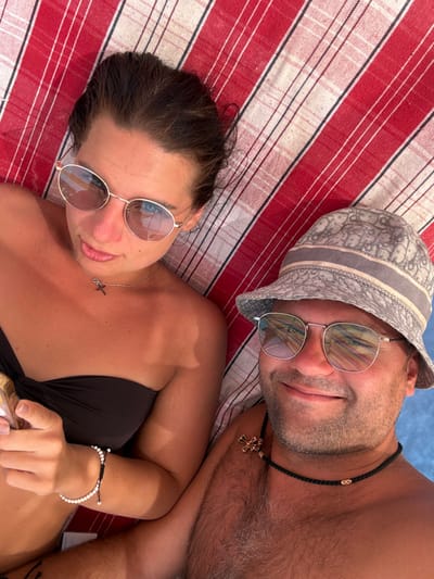 A top-down view depicts two individuals, a female and a male, lying side-by-side on a red and white striped fabric, likely a towel or blanket. The female, positioned on the left, has dark hair, wears a dark strapless top, and round gold-rimmed sunglasses that reflect a blue sky or water. She wears a thin silver chain with a black crucifix pendant and a white beaded bracelet on her right wrist, holding a partially visible gold-colored object. Her expression is a subtle smile directed towards the camera. The male, on the right, wears a light-colored bucket hat with a grey monogram pattern (appearing to be Dior), and silver-rimmed sunglasses also reflecting a blue environment. He wears a dark braided necklace with a prominent gold crucifix pendant and a smaller gold bead. He smiles broadly at the camera. Both individuals exhibit tanned skin. This scene is situated in Bhagatwadi, India.