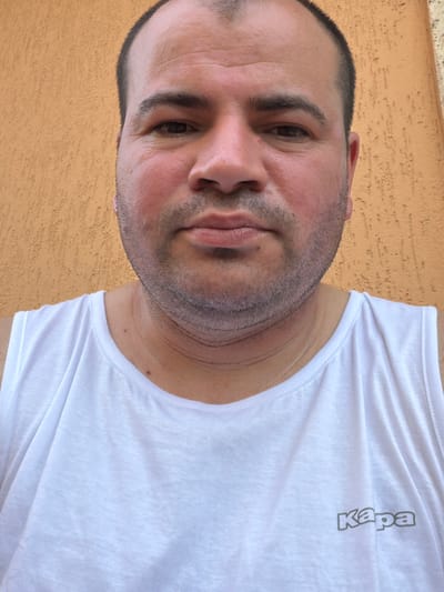 This is a close-up, head-and-shoulders shot of a man wearing a white t-shirt. The image is oriented vertically, with the man's head towards the left and his body extending to the right. He has short, dark hair and a stubbly beard. His skin appears slightly flushed, suggesting he might be outdoors or experiencing exertion. His eyes are dark and looking slightly off-camera. The t-shirt has a printed logo on the upper right chest area, which appears to read "Kapa" in a vertical, gray font. The background is a textured, orange-brown surface, possibly a wall or stucco, consistent with outdoor settings in Italy. The lighting suggests it is daytime, possibly sunny, given the bright illumination on the man's face and t-shirt. No specific event is depicted, but the selfie-style framing and casual attire suggest a personal moment or a casual portrait. The overall impression is one of a candid, informal portrait captured outdoors. The location context provided is Piacenza, Italy.