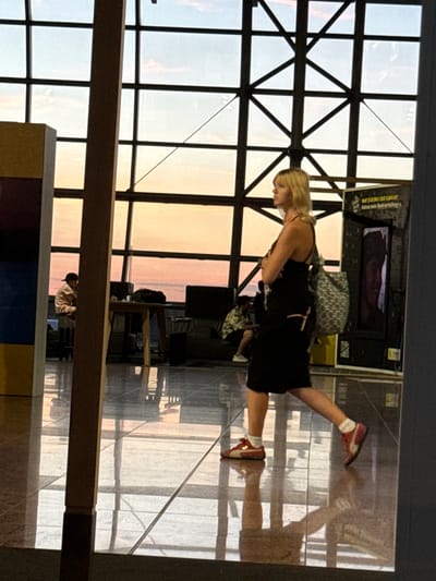 The image depicts a woman walking through a brightly lit interior space with a large window in the background. The window reveals a sunset sky with hues of orange and pink. The woman is wearing a black dress, white socks, and red and white sneakers. She has a patterned backpack slung over her left shoulder. To the left of the woman, a yellow and blue structure is partially visible. In the background, through the window, there are glimpses of a sky with clouds. The floor is highly reflective, mirroring the overhead lighting and structures. Further in the background, a man is seated at a table, and other seating areas are visible. The overall impression is of an airport terminal or a similar public transit hub, given the architecture and the presence of people waiting. The lighting suggests it is late afternoon or early evening.