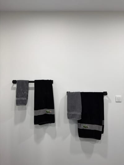 The photograph depicts a section of a stark white wall in an interior setting. Two towel racks, crafted from black metal, are mounted vertically on the wall. Each rack holds two towels: a larger black towel with a gray stripe and a Lacoste logo, and a smaller gray towel. The towels are neatly folded on the racks. A light switch is visible near the top of the frame above the upper towel rack. The wall exhibits a clean, minimalist aesthetic. The location and city are not discernible from the image.