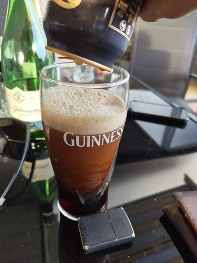 The image shows a glass of Guinness being poured from a bottle. The glass is filled with the characteristic dark liquid and creamy head of the stout. The bottle being poured is a Guinness bottle with its label partially visible. The background includes a bottle of Jägermeister, a Zippo lighter, and a dark, reflective surface.

The location in the city of Schmelz, Germany is not discernible from the given image.